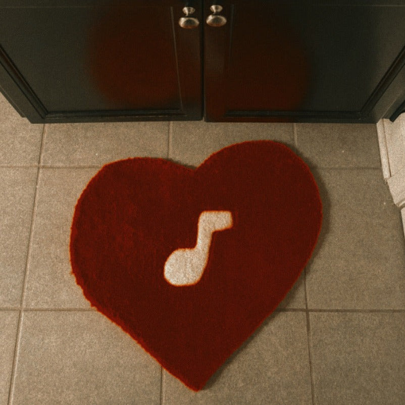 Red heart-shaped tufted rug measuring 21 inches by 21 inches, crafted from 100% acrylic yarn, featuring a white music note design at the center.
